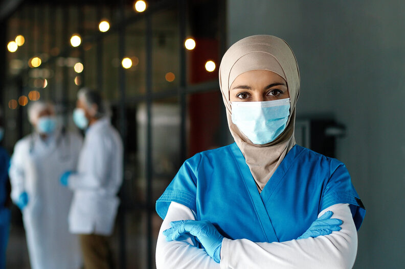A woman wearing scrubs and a mask stands with her arms crossed facing the camera.