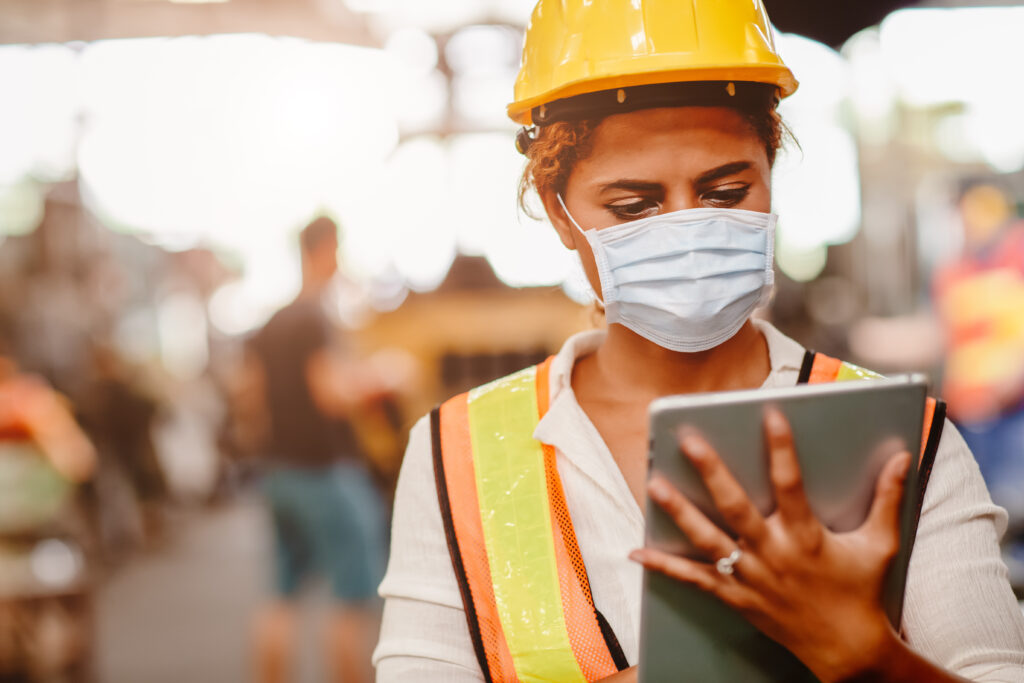 A masked construction workers in a hard hat, reading off of a tablet.